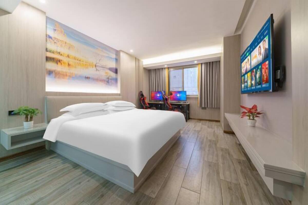 A hotel room with a large bed, a desk with a computer, a TV, and a wall-mounted screen displaying movie options.