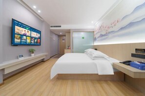 Quarto de hotel com uma televisão de ecrã plano grande fixada na parede, uma cama com roupa de cama branca, uma mesa de cabeceira de madeira e uma secretária com telefone e uma caixa azul.