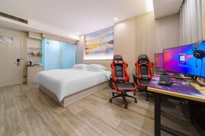 Quarto de hotel com uma cama, duas cadeiras de jogos, uma escrivaninha com computador e fones de ouvido, e uma televisão na parede.