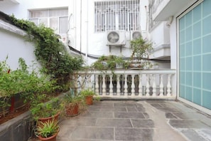 Une cour intérieure agrémentée de plantes en pot, dotée d’un sol en pierre et bordée d’un bâtiment blanc aux fenêtres grillagées et équipé de climatiseurs.