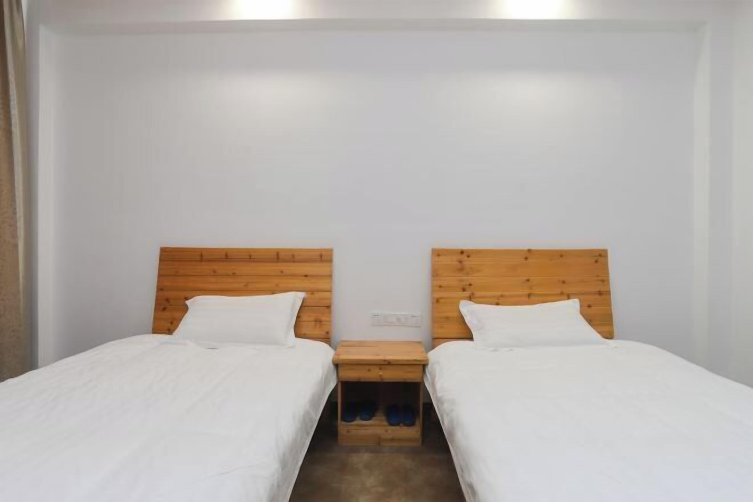 A room with two single beds, wooden headboards, white bedding, and a small wooden nightstand with slippers.