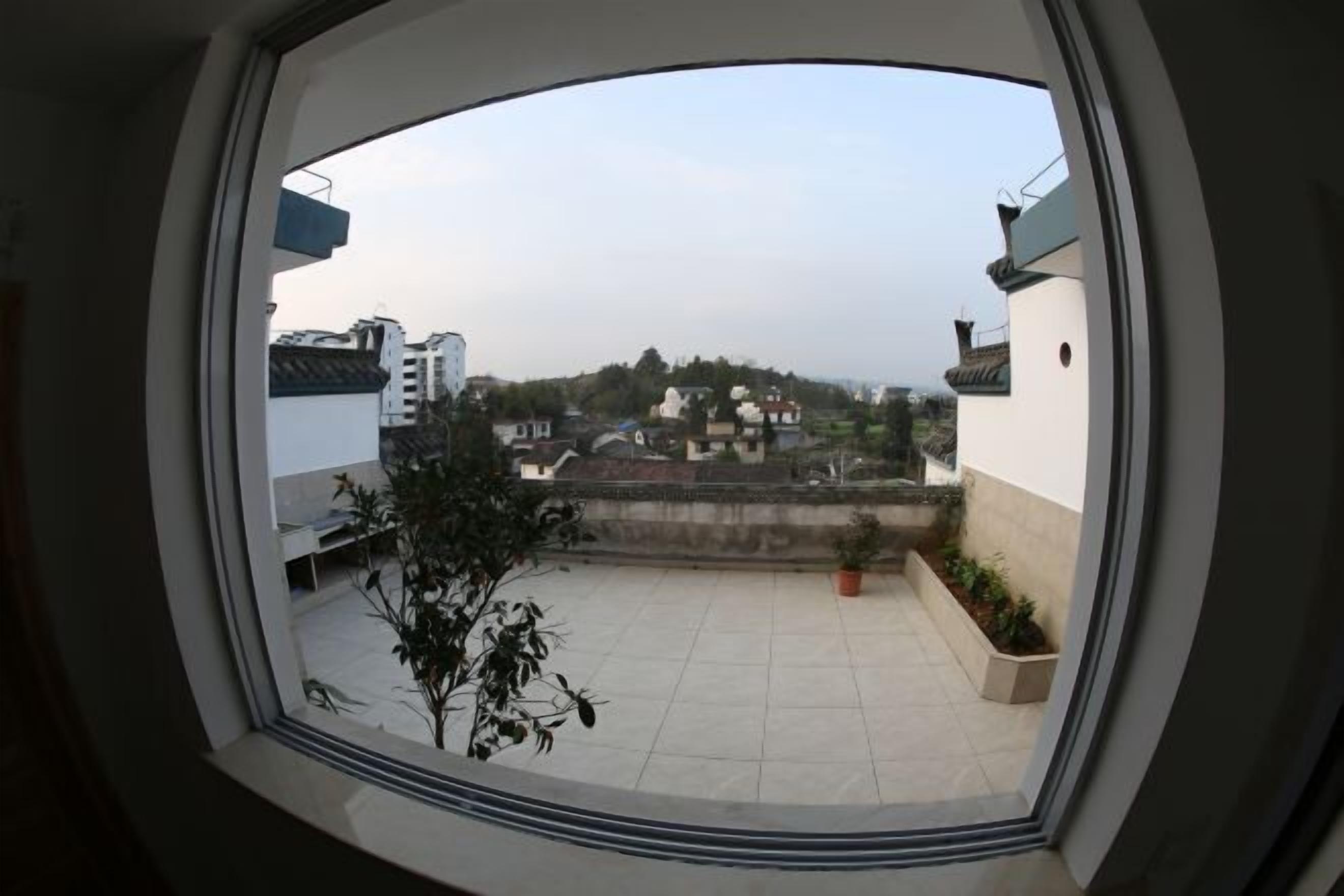 Vista desde una ventana con marco circular, sobre un patio con suelo de baldosas, plantas en macetas y una lejana perspectiva de edificios y una colina.