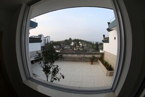 Blick aus einem Fenster mit rundem Rahmen auf einen Innenhof mit Fliesenboden, Pflanzkübeln und eine entfernte Ansicht von Gebäuden und einem Hügel.