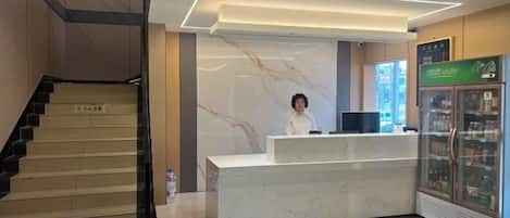 A hotel reception area with a marble reception desk, a staircase, and a refrigerator with beverages.