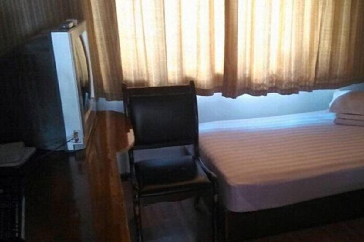 Una cama individual con colchón y almohada blancos, una silla de madera y un televisor sobre un mueble de madera.