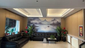 A spacious lobby with a black leather sofa, a coffee table, and potted plants. A large mural of a mountainous landscape is on the wall. - Daguang Hotel (Turpan)