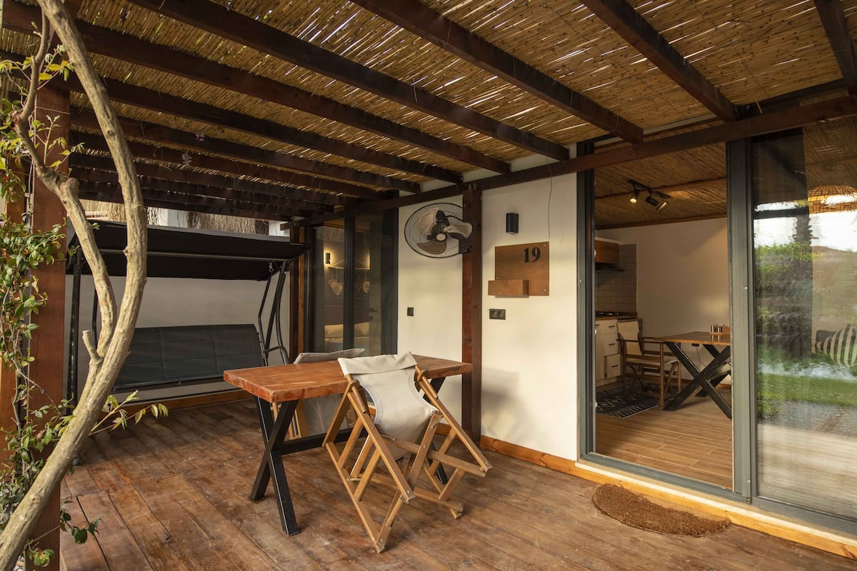 A room with a wooden table, a folding chair, a fan, and a large window.