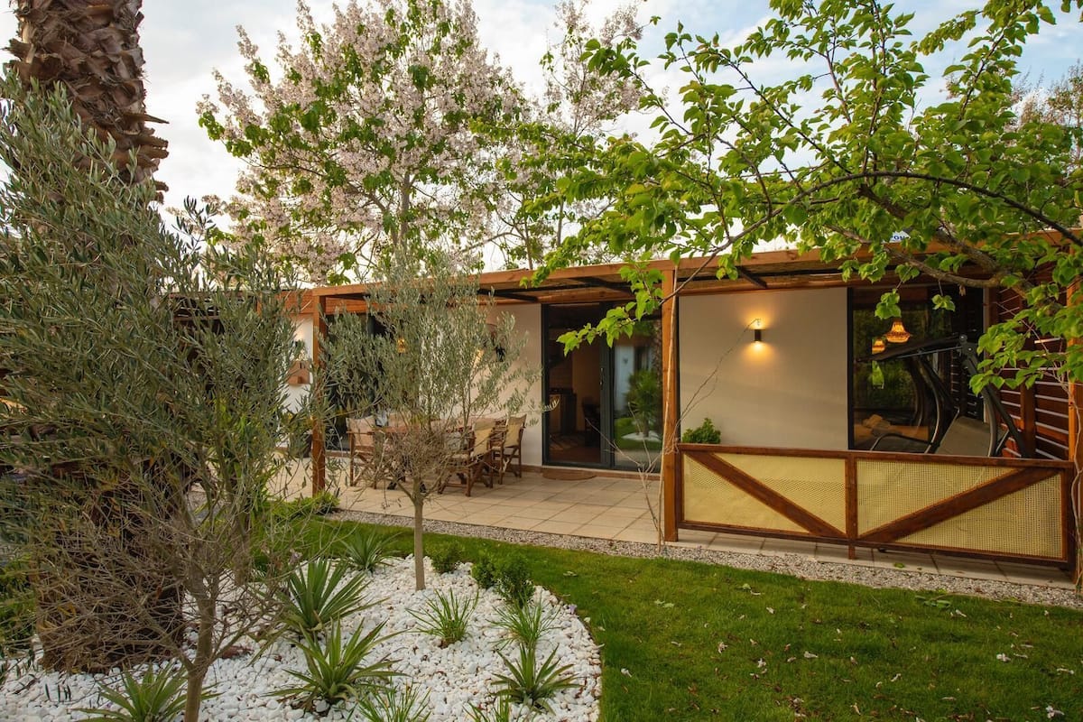 A modern outdoor patio area with a wooden pergola, a seating area with a table and chairs, and a glass enclosure.