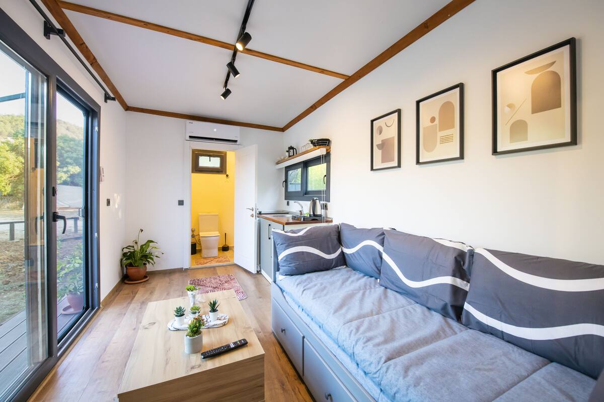 A modern living room with a sofa, a coffee table with plants, and a kitchen area visible through an open door.