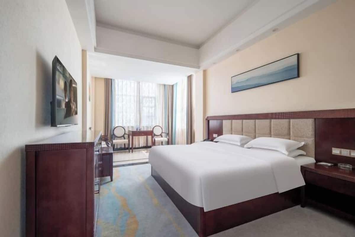 A hotel room with a large bed, a desk, a chair, and a window with curtains.