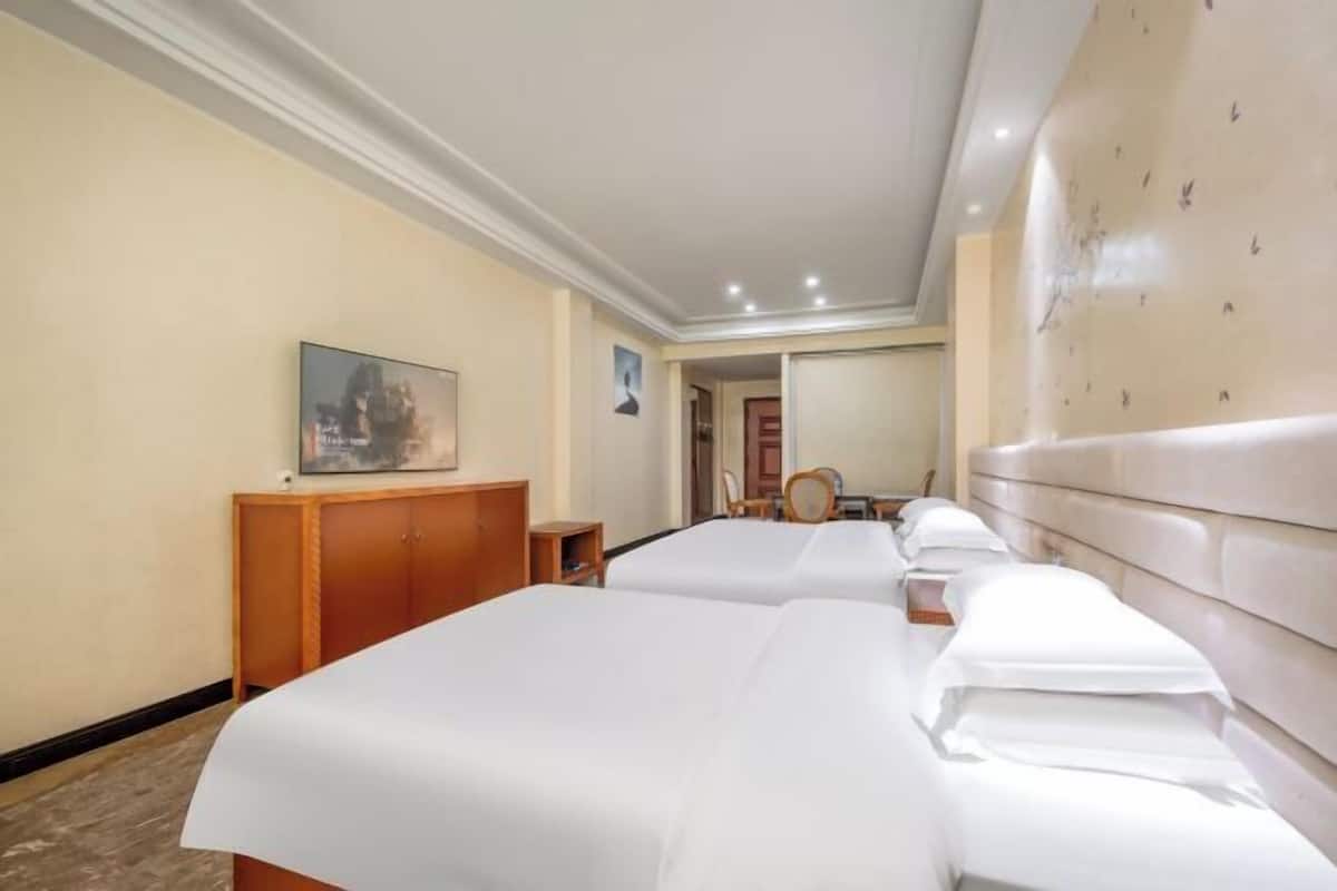 A hotel room with two beds, a wooden wardrobe, a small table, a mirror, and a painting on the wall.