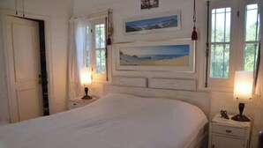 Ein Schlafzimmer mit einem großen Bett, zwei Nachttischen und einem Beistelltisch. Zwei Fenster mit Vorhängen, darüber am Bett eine Wandkunst.
