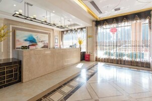 Zona de recepción de un hotel con suelo de mármol, mostrador de recepción y un amplio ventanal con cortinas.