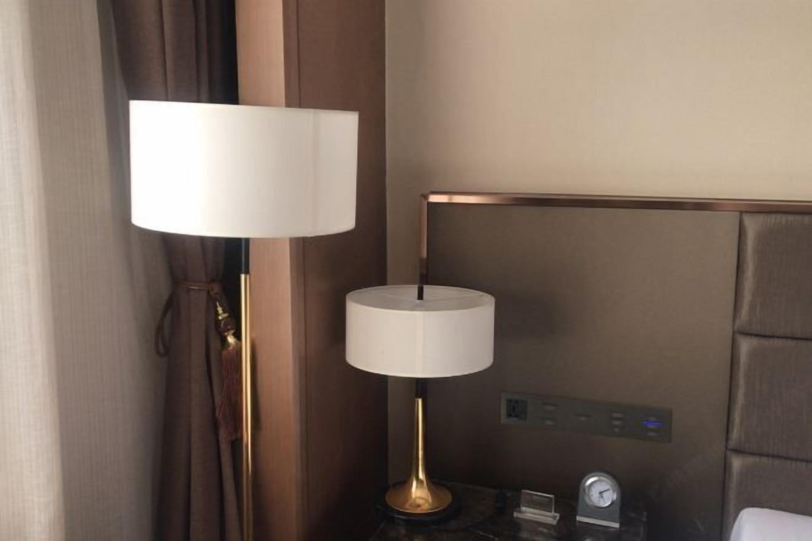 Una camera d'albergo con un letto, un comodino con una lampada, una sveglia e un pannello di controllo a parete.