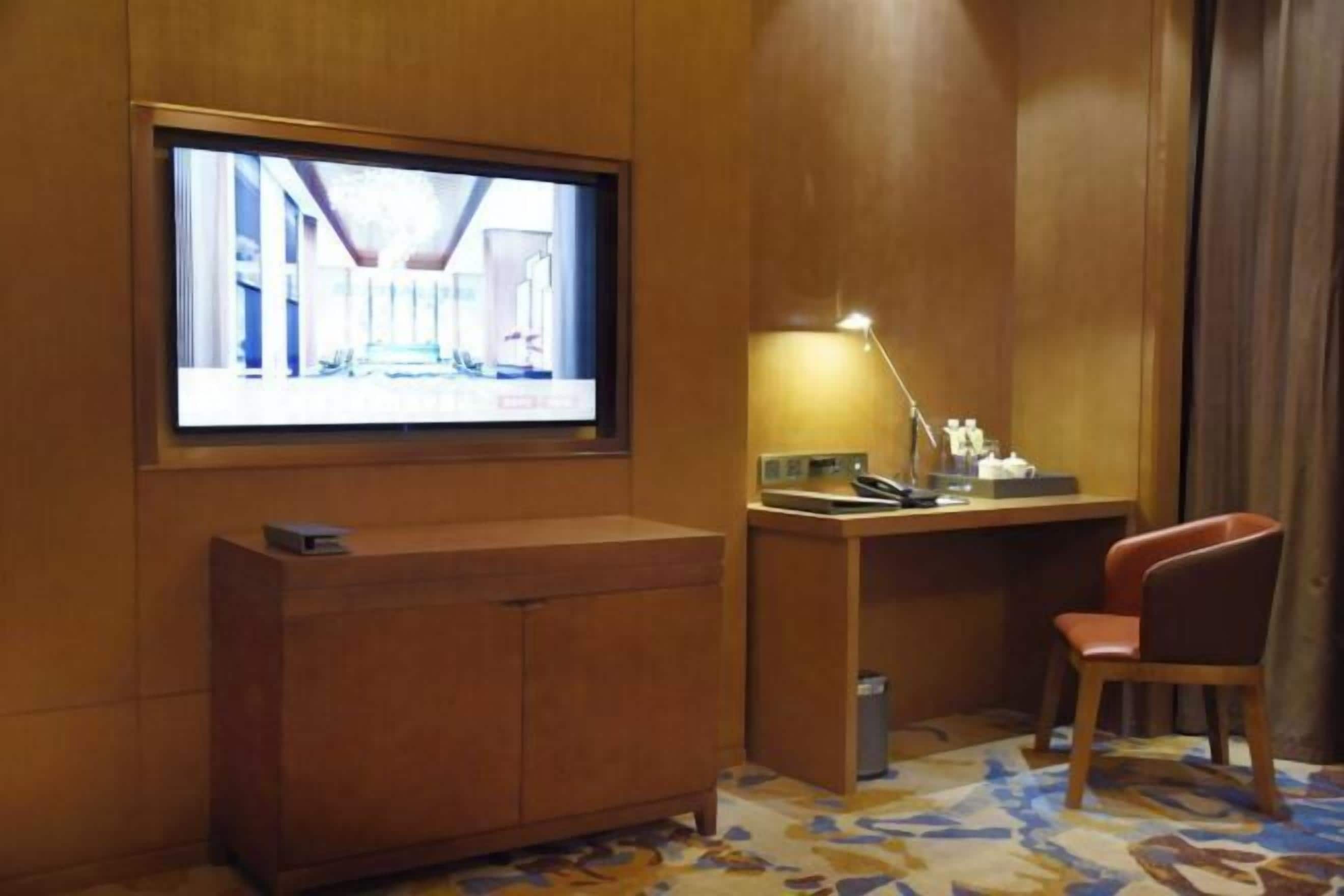 Habitación de hotel con televisión montada en la pared, un escritorio con lámpara, una silla y una alfombra estampada.