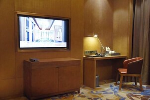 Habitación de hotel con televisión montada en la pared, un escritorio con lámpara, una silla y una alfombra estampada.