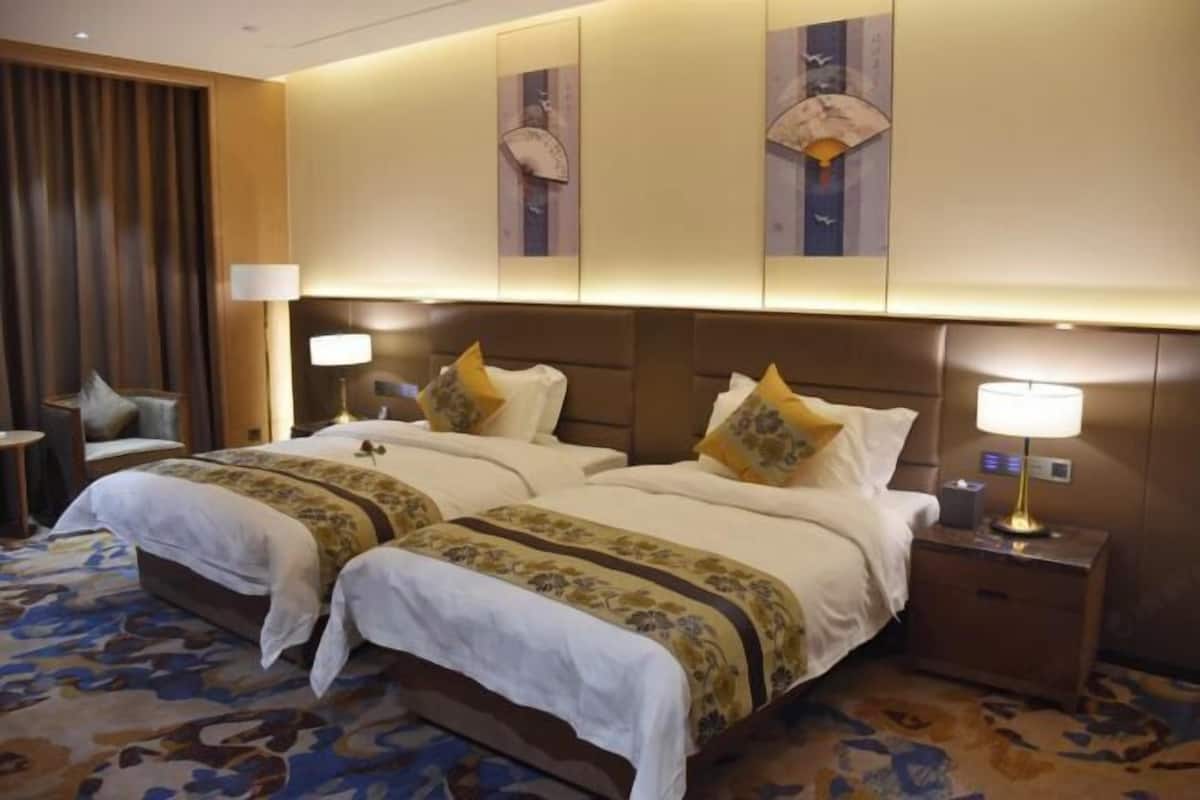 Habitación de hotel con dos camas, un ventanal con cortinas, una mesita de noche con lámpara y un reloj en la pared.