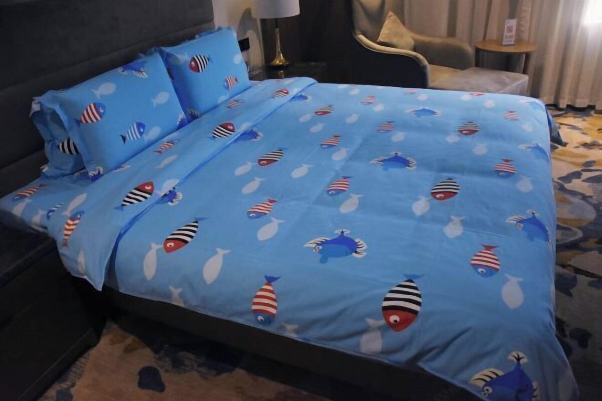 A hotel room with a bed covered in a blue bedspread with fish patterns, a dark headboard, a small round table, and a chair.
