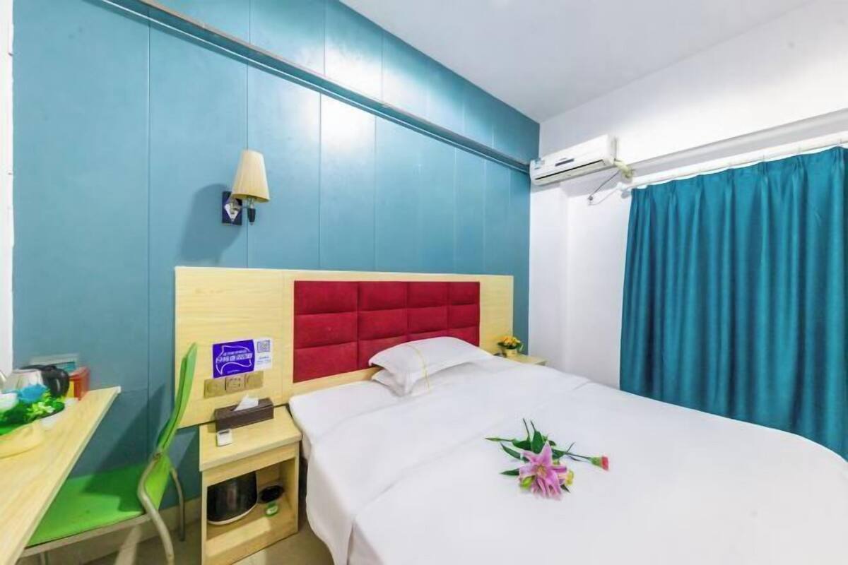 Une chambre d’hôtel comprenant un lit, un bureau, une chaise, une table de chevet, une lampe, un climatiseur fixé au mur et un rideau.
