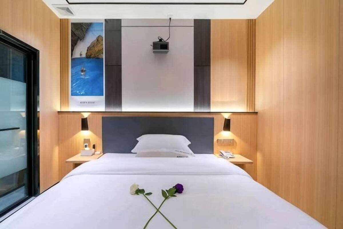 Une chambre d’hôtel avec un lit, des tables de chevet, une petite table, une lampe fixée au mur et une affiche accrochée au mur.