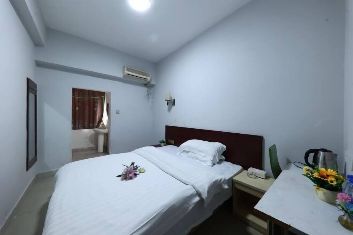 Une chambre d’hôtel comprenant un lit, une table de chevet, un bureau, un climatiseur, une fenêtre avec des rideaux et un luminaire fixé au mur.