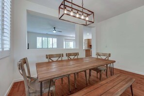 A dining area with a wooden table and chairs, a hanging light fixture, and a view into the kitchen.