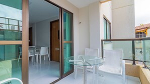 Un balcón con una mesa de vidrio y sillas de mimbre blanco, una puerta corrediza de vidrio que da a un espacio interior con una mesa y sillas, y vistas de un edificio con techo de tejas.