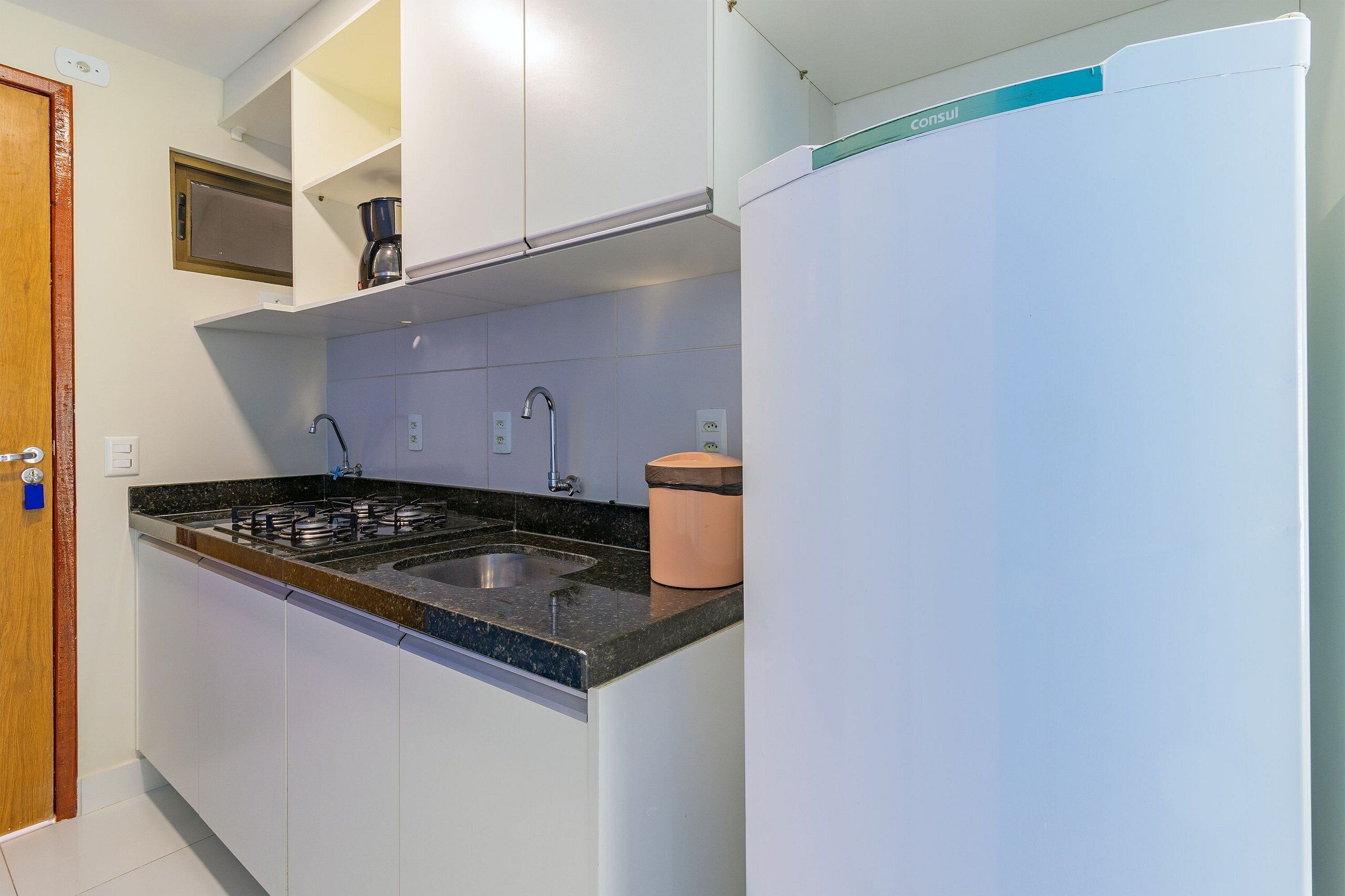 Una cocina con armarios blancos, encimera negra, lavabo, cafetera y refrigerador.