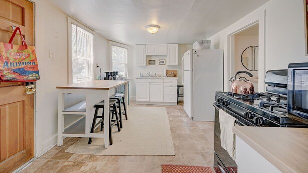 A compact kitchen with white cabinets, a stove, and a refrigerator. There is a dining area with stools and a small table.