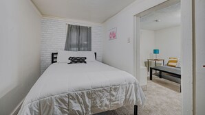 A bedroom with a bed, a white brick wall, a window with curtains, a desk with a lamp, and a chair.