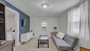 A living room with a grey sofa, a white TV stand, a small table, and a window with white curtains.