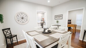 A dining area with a wooden table, white chairs, and a decorative wall piece. - Private Patio W/ Grill + Game Room - Sleeps 10! (Wichita)