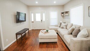 A living room with hardwood flooring, a beige sectional sofa, a bench, and a flat-screen TV mounted on the wall. - Private Patio W/ Grill + Game Room - Sleeps 10! (Wichita)