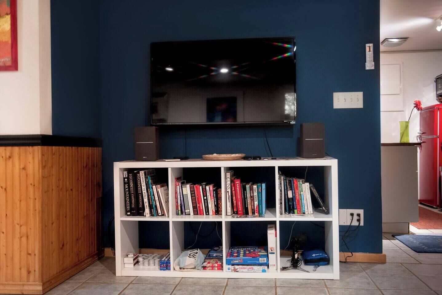 A room with a TV mounted on the wall, a white bookshelf with books and electronic devices, and a wooden cabinet.