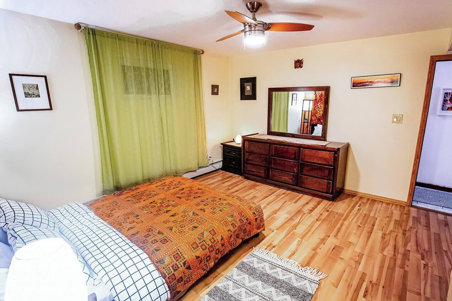Ein Schlafzimmer mit Holzboden, einem Bett mit gemusterten Betttextilien, einer Kommode, einem Deckenventilator und einem Spiegel an der Wand.