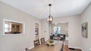 Comedor con mesa y sillas de madera, lámpara colgante y vista a una cocina y sala de estar.