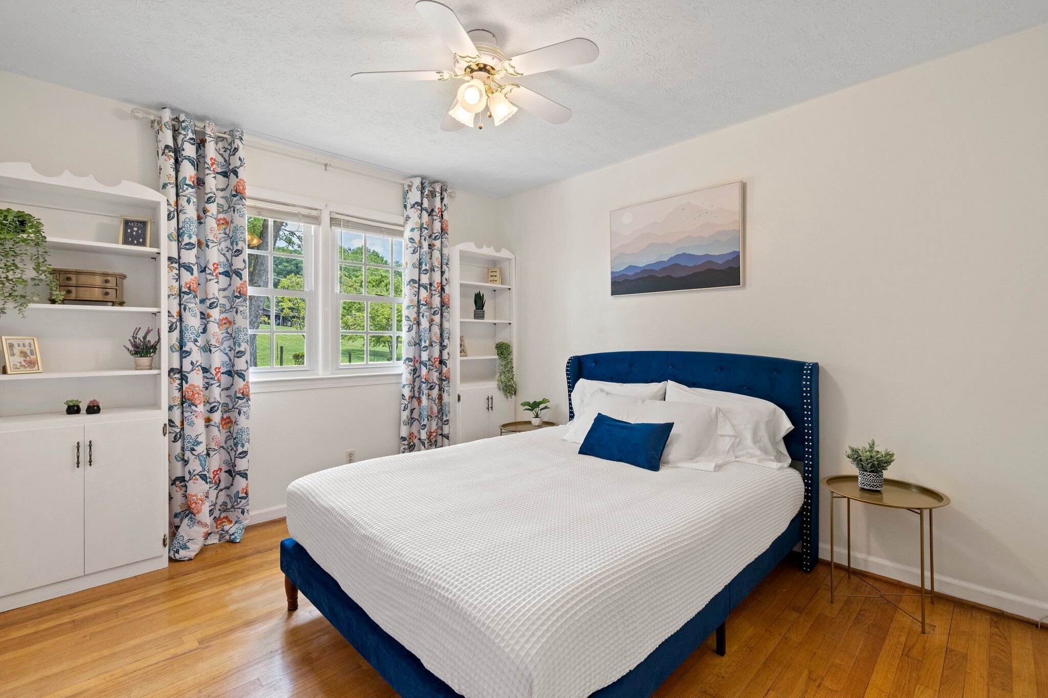 A bedroom with a bed, a ceiling fan, a window with curtains, a bedside table, and a wall-mounted shelf.