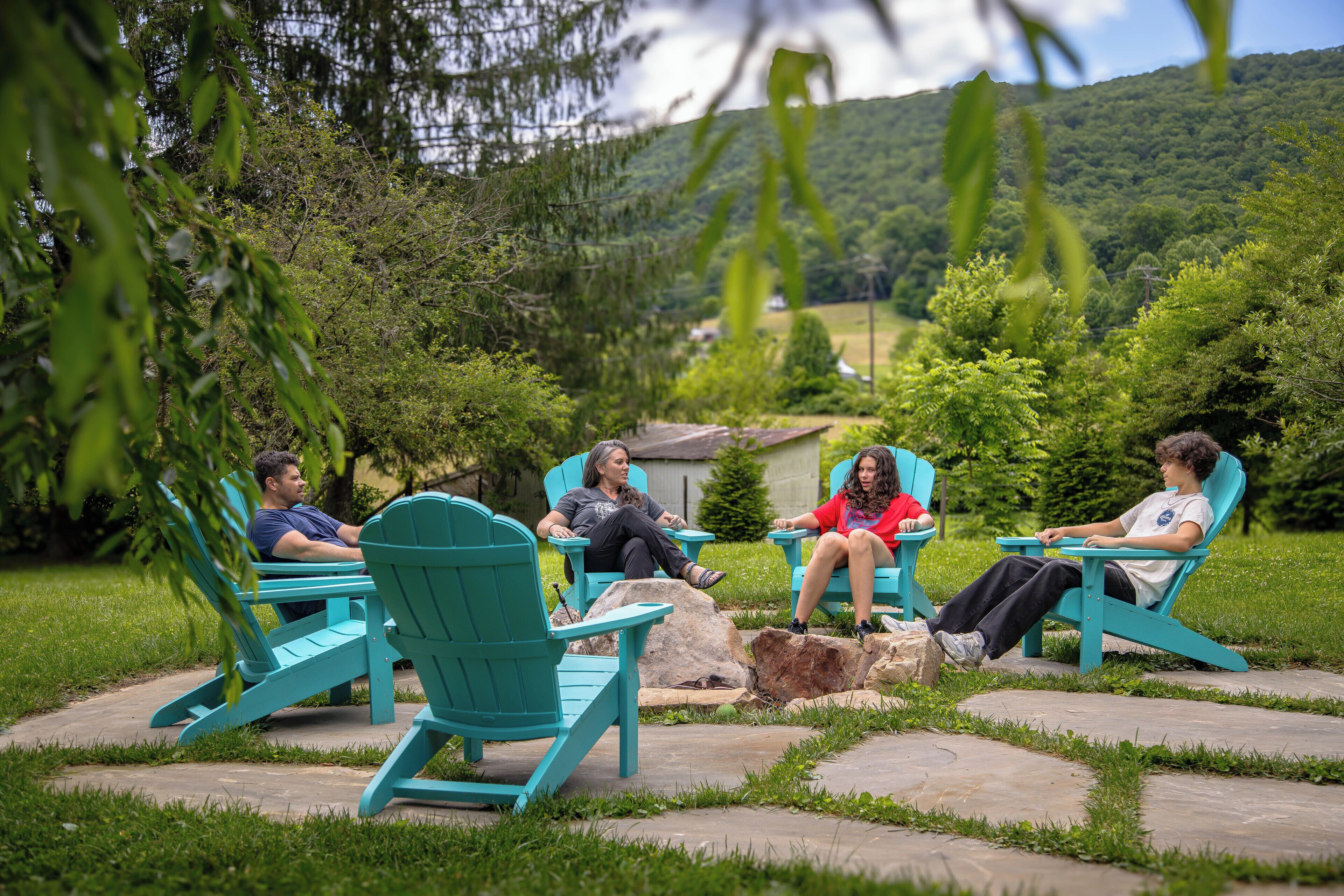 Cuatro personas sentadas en sillas Adirondack turquesa en un jardín con un patio de piedra, rodeado de abundante vegetación y un paisaje montañoso al fondo.