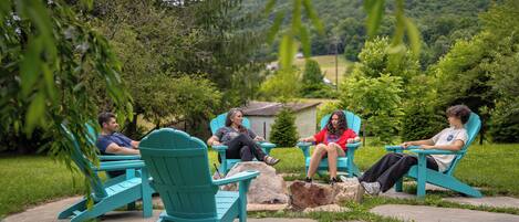 Cuatro personas sentadas en sillas Adirondack turquesa en un jardín con un patio de piedra, rodeado de abundante vegetación y un paisaje montañoso al fondo.