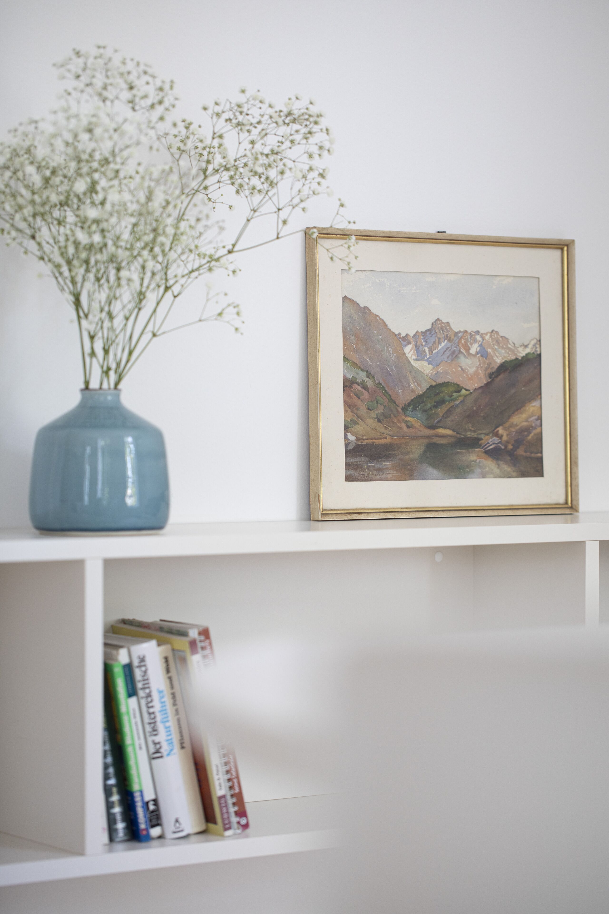 Ein gerahmtes Landschaftsbild auf einem weißen Regal, eine blaue Vase mit weißen Blumen und eine Reihe Bücher.