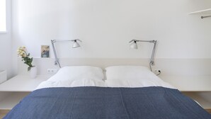 Ett modernt sovrum med en säng, sänglampor, ett nattduksbord, en vas med blommor och en vägghylla.