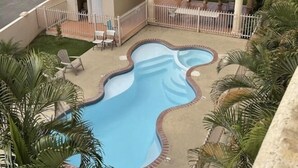 Une piscine à bordures ondulées, entourée d’une clôture et de palmiers. On y trouve deux petits bâtiments aux toits recouverts de tuiles rouges et un espace de repos abrité.