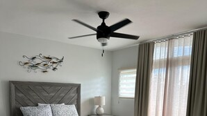 Une chambre à coucher avec un lit en bois, un ventilateur de plafond, une table de chevet, une lampe et une fenêtre avec des rideaux.