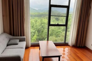 Quarto com sofá cinzento, mesa de madeira e uma grande janela com vista para uma paisagem arborizada.