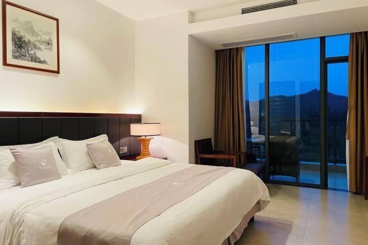A hotel room with a bed, a desk, a chair, a lamp, and a large window with curtains.