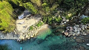 Vista aérea de una playa con agua cristalina, rocas y una pequeña cabaña.