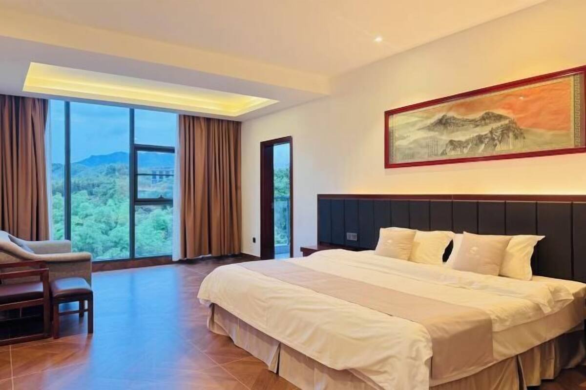 A hotel room with a large bed, a chair, a painting, and a view of the outdoors.
