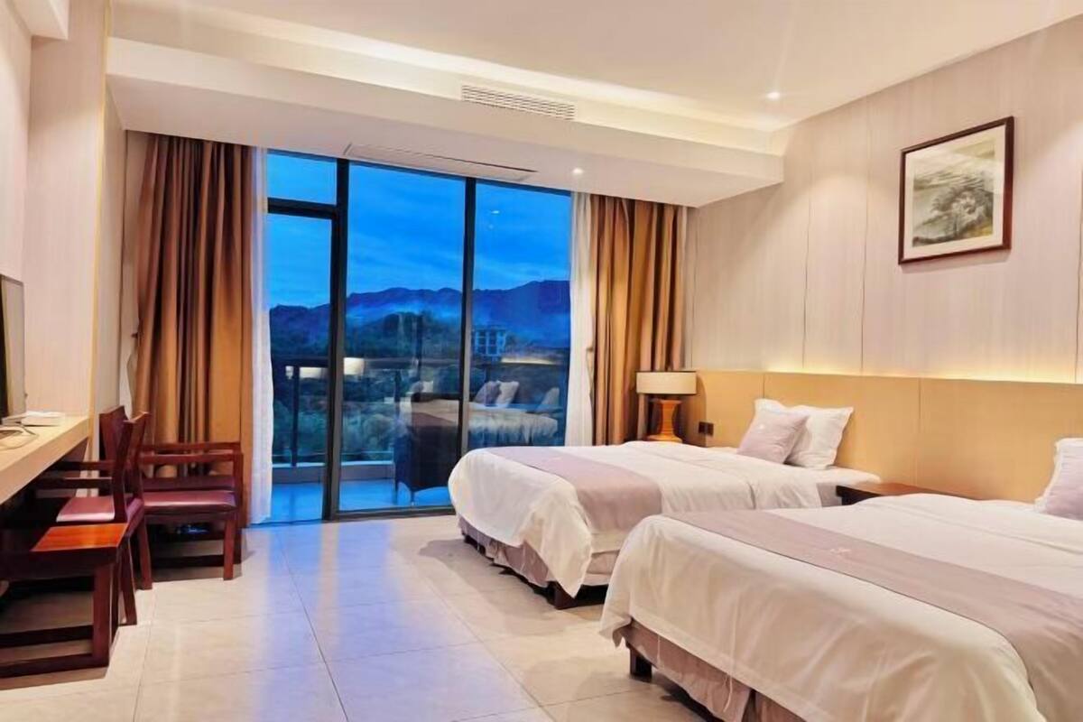 A hotel room with two beds, a desk, chairs, a large window with a view, and a painting on the wall.