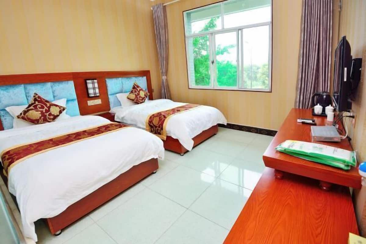 A hotel room with two beds, a wooden desk, a television, and a window with a view of greenery.