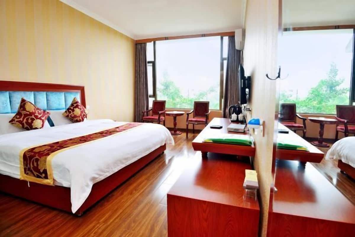 A hotel room with a bed, a desk, a chair, and a view of greenery outside.